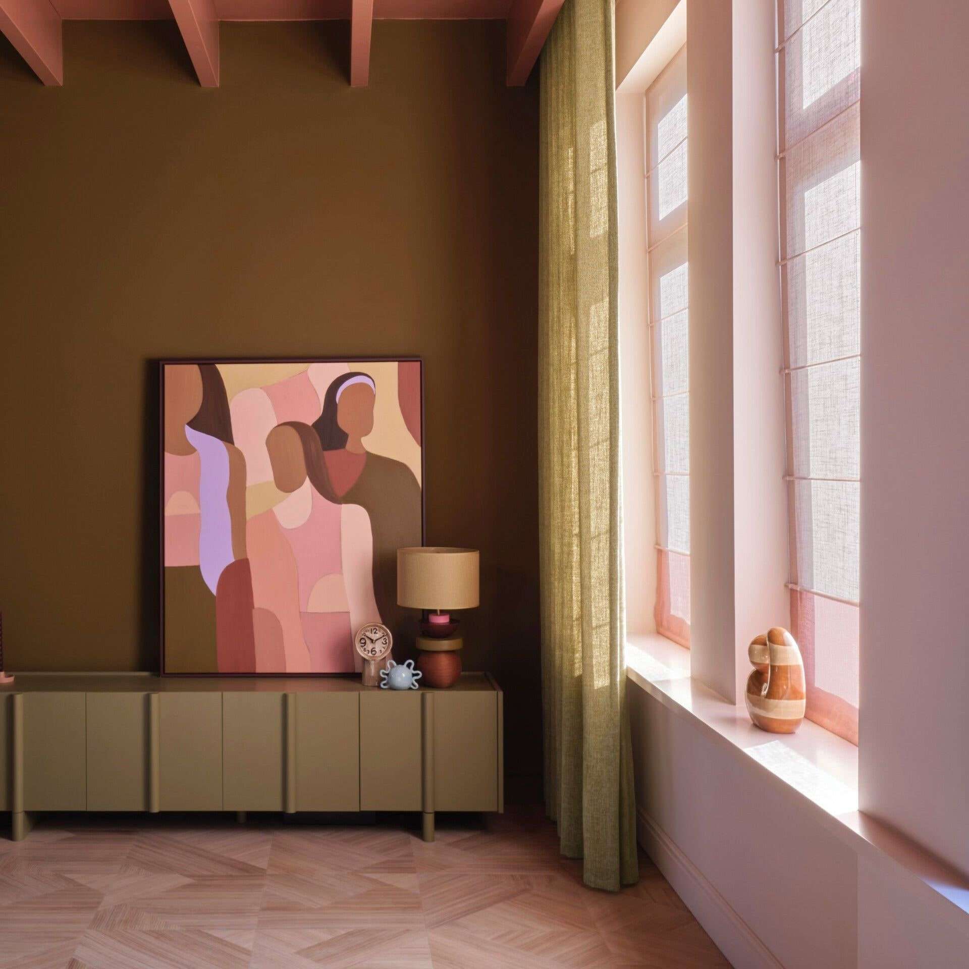Een kamer met een olijfgroene wand, een laag dressoir met een schilderij en een lamp, een linnen gordijn en roze vouwgordijnen voor hoge ramen, op een lichte houten vloer